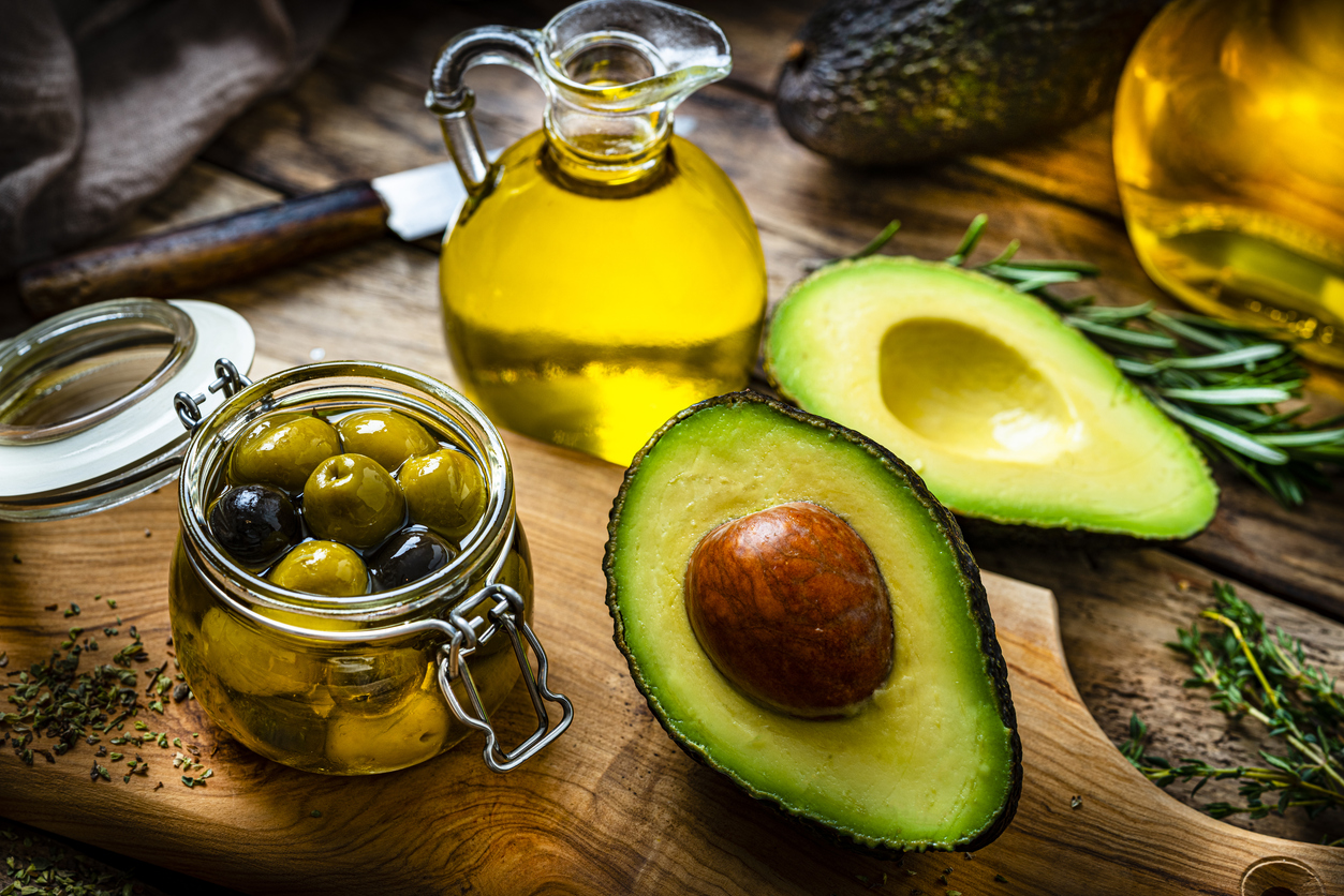 Vegan food: high angle view of extra virgin olive oil and avocado oil shot on rustic wooden table. Sliced organic avocado and a glass container with olives complete the composition. Predominant colors are yellow and green. High resolution 42Mp studio digital capture taken with Sony A7rII and Sony FE 90mm f2.8 macro G OSS lens