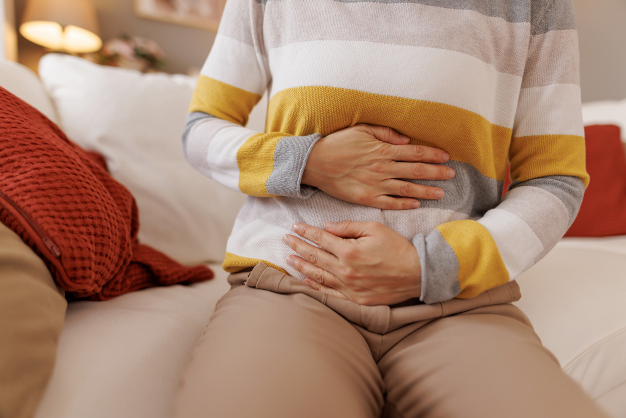 Mature woman experiencing stomach or abdominal pain while sitting on a comfortable sofa in her living room, a common symptom of various digestive issues